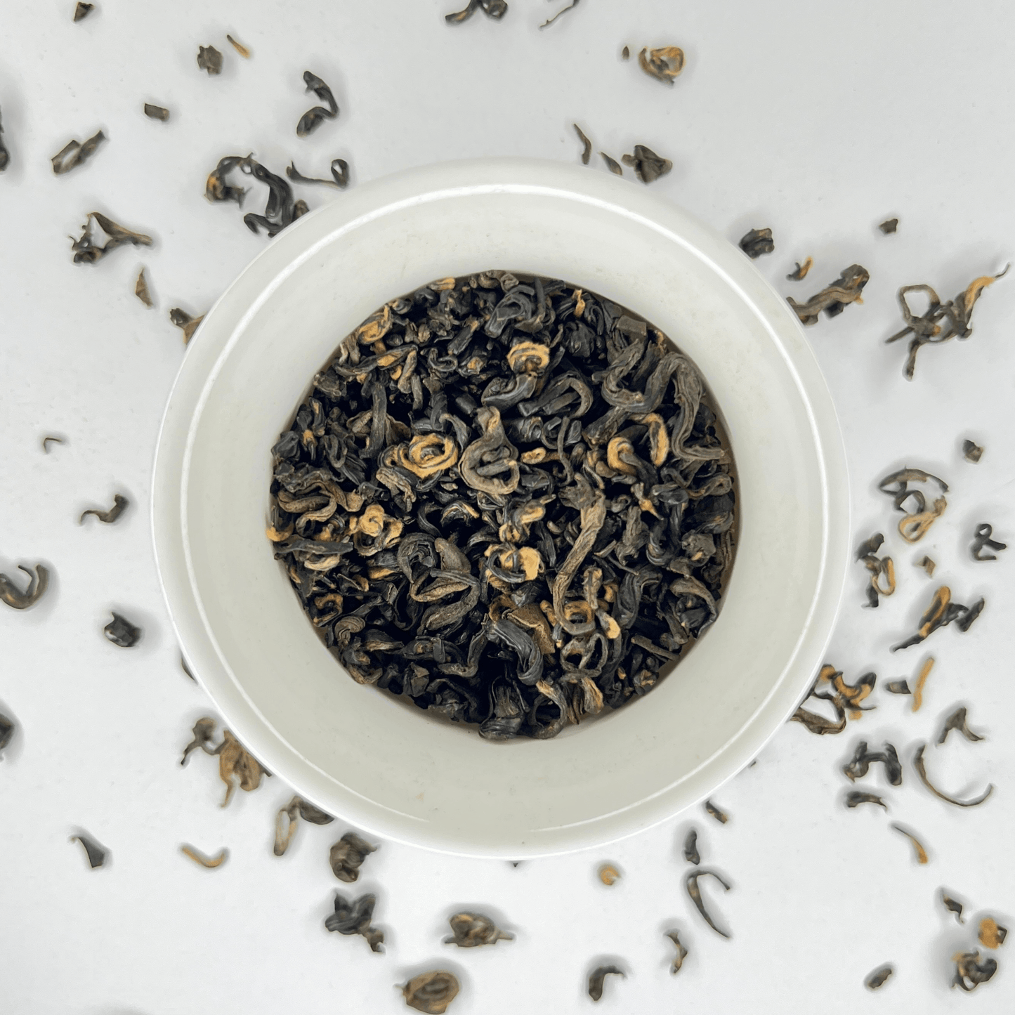 White bowl filled with black tea leaves on a light gray background with scattered tea leaves.