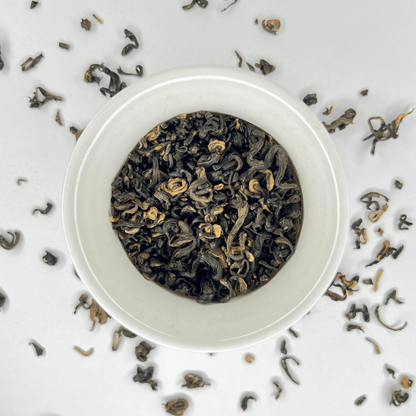 White bowl filled with black tea leaves on a light gray background with scattered tea leaves.